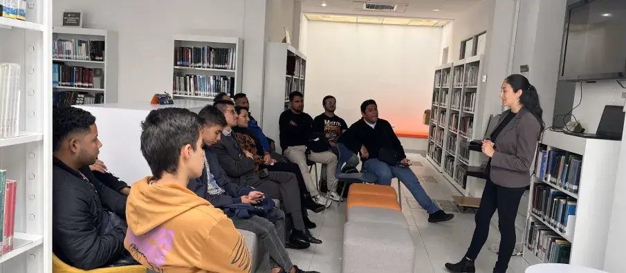 Varias personas en una reunión, con un estante de libros al fondo.