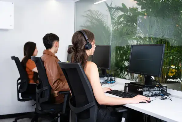 Tres personas sentadas frente a computadoras, una de ellas con auriculares, en una sala con pared de vidrio y plantas al fondo.